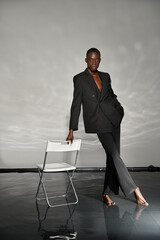 tempting african american man in sexy black suit posing next to chair on gray watery backdrop