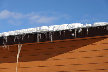 Many melting icicles with fall down drops hangs down close-up on the flat roof with snow against...