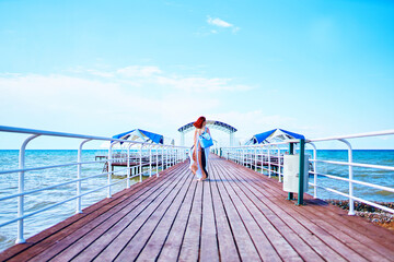 Amidst the resort pier and the tranquil sea, a woman with beautiful red hair, dressed in a summer ensemble and carrying a backpack, takes a leisurely stroll.