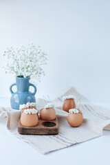 On a linen napkin there is a wooden egg cup with brown Easter eggs with flower wreaths and bunny ears. In the background there is a blue vase with gypsophila flowers. White background