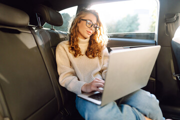 Beautiful woman using laptop while sitting on a backseat of a car. Remote work. Concept of travel, technology, internet.