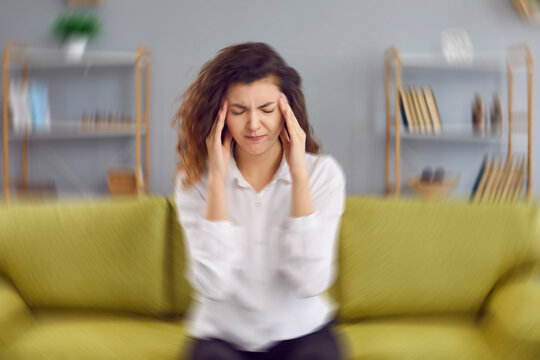 Woman feels dizzy and sits down on the couch. Young woman sitting on the sofa at home, feeling pain and a spinning sensation in her head. Concept of headache, vertigo, health problem, brain tumor