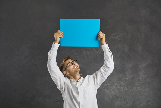 Happy smiling Caucasian man looks at a blank blue sheet of paper he is holding over his head. Man makes an announcement, votes or expresses his position, standing on a gray background. Promotion.