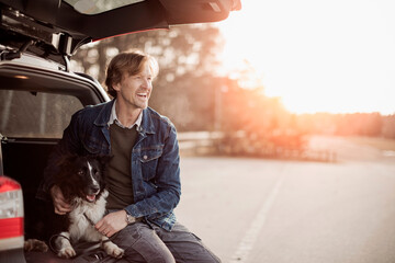 Mature man sitting in a car trunk with his dog at the park - obrazy, fototapety, plakaty