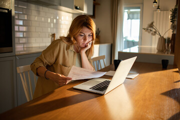 Worried woman reviewing documents with laptop at kitchen table - obrazy, fototapety, plakaty