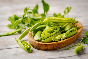 Fresh green peas in wooden bowl with pods and leaves on white wooden table, healthy green vegetable or legume