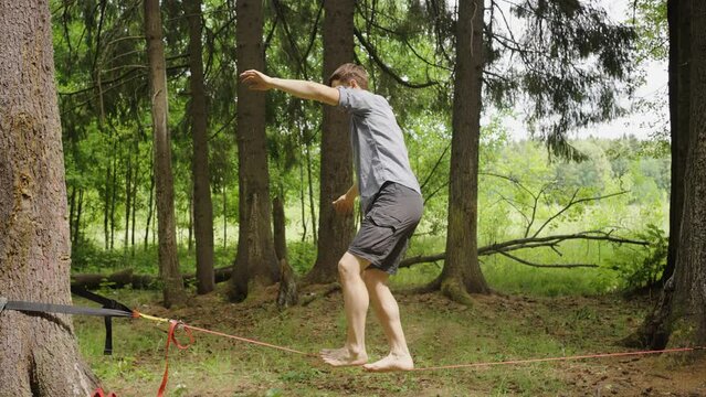 A man balances barefoot on a slackline, arms outstretched for stability, amidst the tranquil backdrop of a dense forest. An individual practices slacklining between trees in a lush forest, a test of