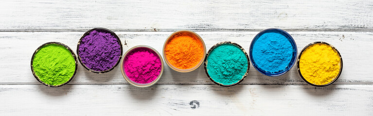 Colored Holi powder in bowls in a row on a white wooden table. Top view, banner. Traditional...