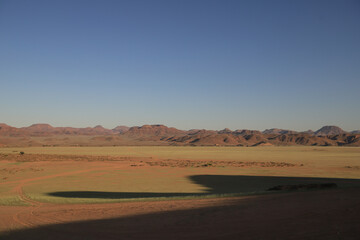 Fototapeta na wymiar red desert sand plaine with bright green fresh grass growing, red mountain range in the background