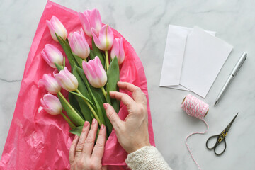 Fototapeta na wymiar Female hands arranging pastel pink tulips bouquet on a florist table with blank card