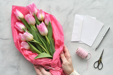Female hands arranging pastel pink tulips bouquet on a florist table with blank card