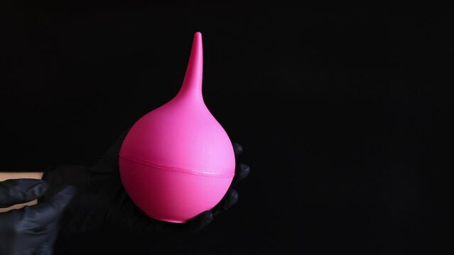 A hand in a black latex glove holds a large pink enema on a dark background. Medical device. Pear-shaped enema, body cleansing concept
