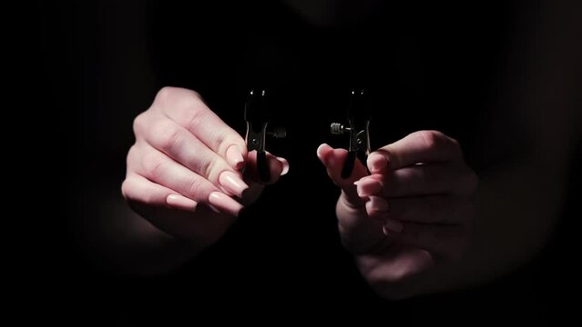 Two metal BDSM nipple clamps in female hands on a black background. Products for intimate stores, sex toys, adult gifts for couples
