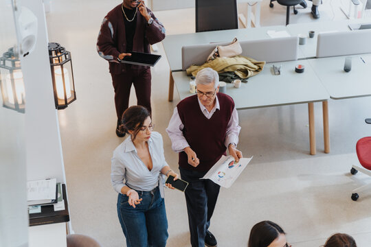 An Elevated View Of A Modern Office Space With Diverse Coworkers Engaging In Work Activities. Some Are Walking And Discussing Business While Another Is On The Phone.