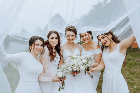 Wedding photography in nature. A brunette bride and her girlfriends pose under a veil, holding bouquets