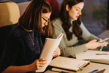 Two businesswomen engrossed in work with documents and digital devices during late hours in a...