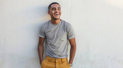 A joyous man in a gray t-shirt and yellow pants is standing against a white wall, laughing with his...
