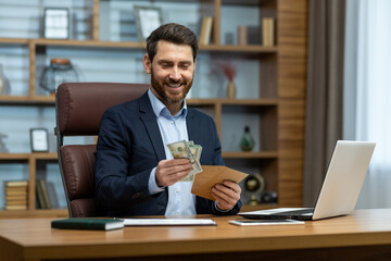 Photo of a happy young businessman sitting at a desk in the office and holding an envelope with...