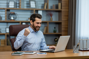 Fototapeta na wymiar Happy young business man sitting in the office at the desk with a notebook and rejoices at the success, looking at the window, showing a victory gesture with his hand