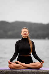 Young fitness woman sits on a caremat in the lotus position on a blurred background of the embankment
