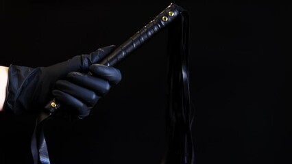 A hand in a black latex glove holds a whip against a dark background. Leather whip for spanking isolated on black. Sex toy for intimacy. Sexual slavery