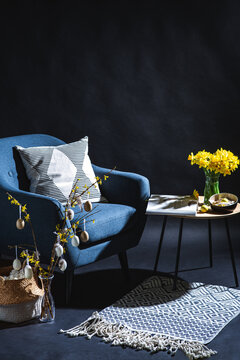 interior, holidays and home decor concept - modern blue chair with pillow, basket and easter eggs hanging on willow branches on table in dark room