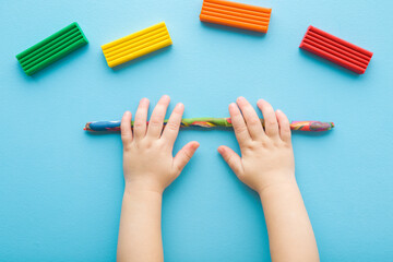 Baby boy hands rolling colorful stick from modeling clay on light blue table background. Pastel...