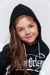 Close-up portrait of little stylish smiling kid girl wearing a black hoodie on white studio background