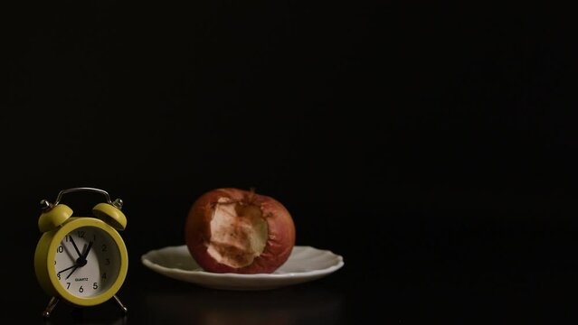 A yellow alarm clock shows the time against the background of a rotten apple on a saucer, black background. Passage of time, aging concept