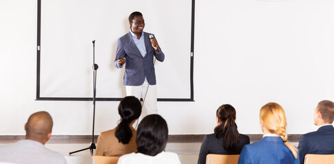 Smiling african american lecturer standing with microphone on stage in conference room, speaking to...