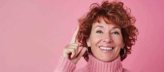 Obraz na płótnie Canvas A middle-aged woman with curly hair wearing a pink turtleneck sweater is holding a toothbrush in her hand, smiling warmly in front of a pink background. She appears to be getting ready for her daily
