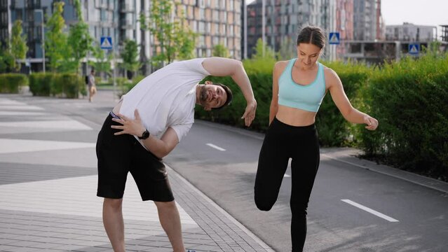 A young positive sportive couple does morning exercises before workout in the city park area among modern residential highrise buildings. A smiling girl and a guy make a high five gesture and start