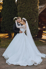 Stylish, young groom and beautiful bride in a long white dress and a long veil with a bouquet in their hands, hugging in the park in the autumn nature. Wedding portrait of newlyweds.