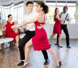 Graceful duet, expressive young Hispanic man in formal wear and slender woman in vibrant red dress, engrossed in passionate tango during group dance class in choreographic studio..