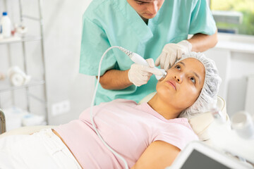 Fototapeta premium Young male cosmetologist performs hardware facial ultrasound procedure to young female patient