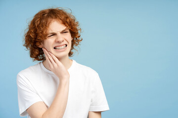 Curly red haired teen boy with braces, holding his cheek in pain, against a blue background