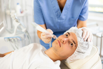 Young woman cosmetologist performs facial cleansing procedure with apparatus young male patient