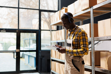 Warehouse employee wearing headphones, writing in clipboard and singing along. African american...