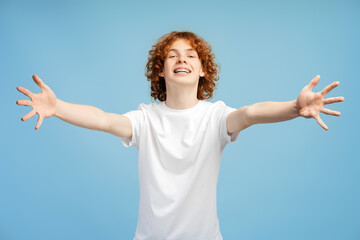 Red haired teenager male with braces, extending arms in welcoming hug pose on blue background - obrazy, fototapety, plakaty