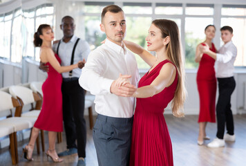 Concentrated elegant young adult man enjoying slow foxtrot with stylish female partner in red dress...