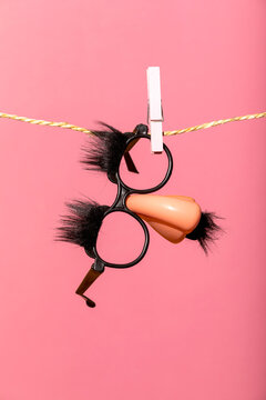 Generative AI illustration of funny novelty glasses with fake nose and eyebrows hanging on a clothespin and string against a pink background