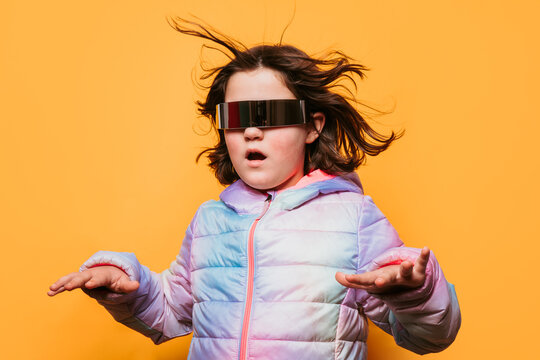 Astonished girl with windblown hair wearing futuristic glasses and a pastel coat, with arms outstretched on an orange background