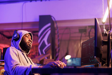 Relaxed african american criminal in hood hacking computer server while listening to music in headphones. Man hacker in earphones wearing hood planning phishing online scam