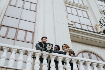 A diverse group of three friends casually collaborates using a laptop on a classic building's balcony, portraying teamwork and friendship.