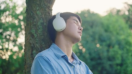 Tired asian young working man sits down under tree, relaxing fall as sleep while listening with white headphone. Digital life entertainment. Domestic lifestyle. Leisure Concept. Relief working stress