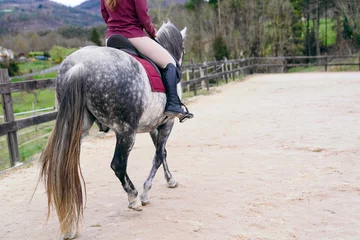 Fotobehang Unrecognizable A young woman enjoys horseback riding on a dappled grey horse in a sandy equestrian arena. © Koldo_Studio