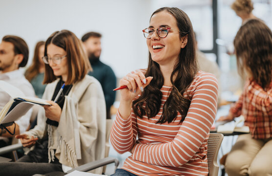 A cheerful young woman wearing glasses and a striped shirt participates in a casual group setting, possibly a classroom or collaborative meeting.