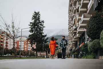 A stylish couple walks along a city sidewalk with a suitcase, showcasing urban travel and a modern lifestyle against an apartment backdrop.