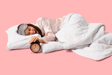 Angry young woman with blanket turning alarm clock off on pink background