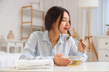 Young Asian woman preparing steam inhalation with herbs at table in bedroom